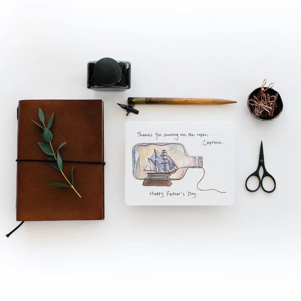 A sailing ship in a bottle on a white background with the words: "Thanks Father's Day" written on it. This card i s on a white table in between a pair of scissors and a diary.
