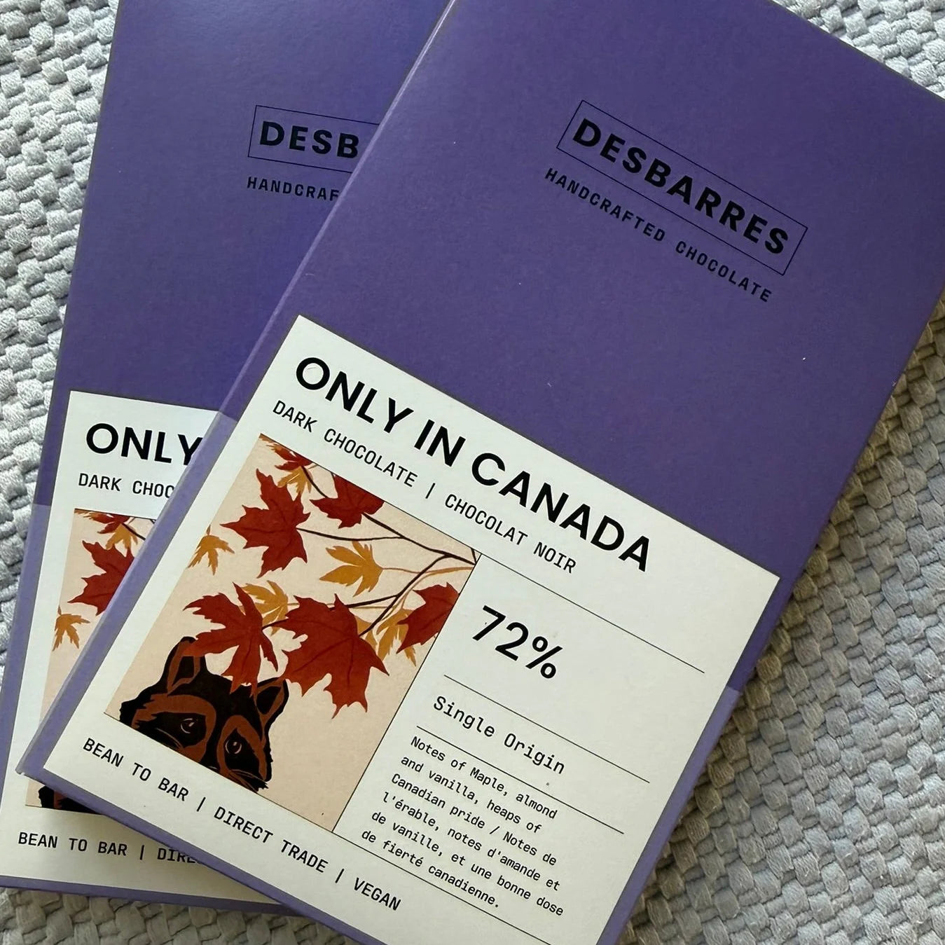 A close-up, overhead view showcases two bars of "Desbarres" handcrafted chocolate, each elegantly packaged in a deep purple cardboard sleeve, with one bar angled slightly over the other on a textured grey surface.
The key subjects are the two chocolate bars. Both feature a minimalist design with the brand name "DESBARRES" in a modern, sans-serif font within a rectangular frame, followed by "HANDCRAFTED CHOCOLATE." The prominent feature is the label on the front of the lower bar, which reads "ONLY IN CANADA"