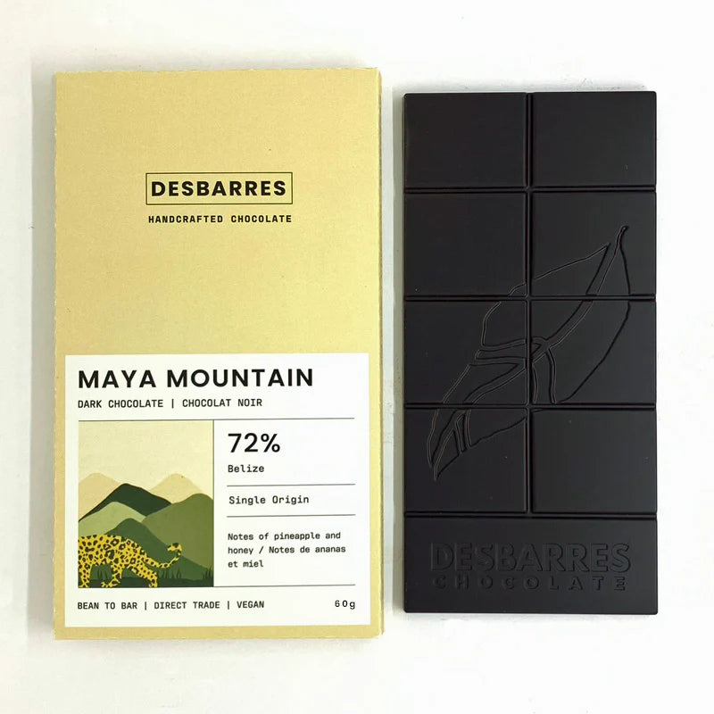 This image displays a bar of Desbarres Maya Mountain 72% dark chocolate and its accompanying pale yellow packaging, presented side-by-side against a clean white background. 
The primary subject is a bar of dark chocolate, presented as a rectangular slab divided into eight individual squares. The chocolate is a deep, rich black, suggesting a high cocoa content. Embossed on its surface is a stylized depiction of a cacao pod or leaf, a common motif for chocolate products. Below the embossed design, the brand n