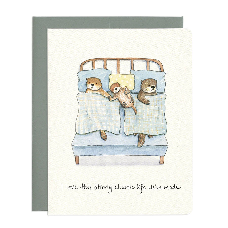The image features a watercolor illustration of a family of otters in bed. The card itself is white with a textured surface. Under the card is a grey envelope.

The central scene depicts a bed with a light blue mattress and a wooden headboard with simple horizontal slats. Two adult otters and one smaller otter are nestled under light blue blankets with a subtle checkered pattern. 
Below the illustration, handwritten in black ink, is the text "I love this otterly chaotic life we've made." 